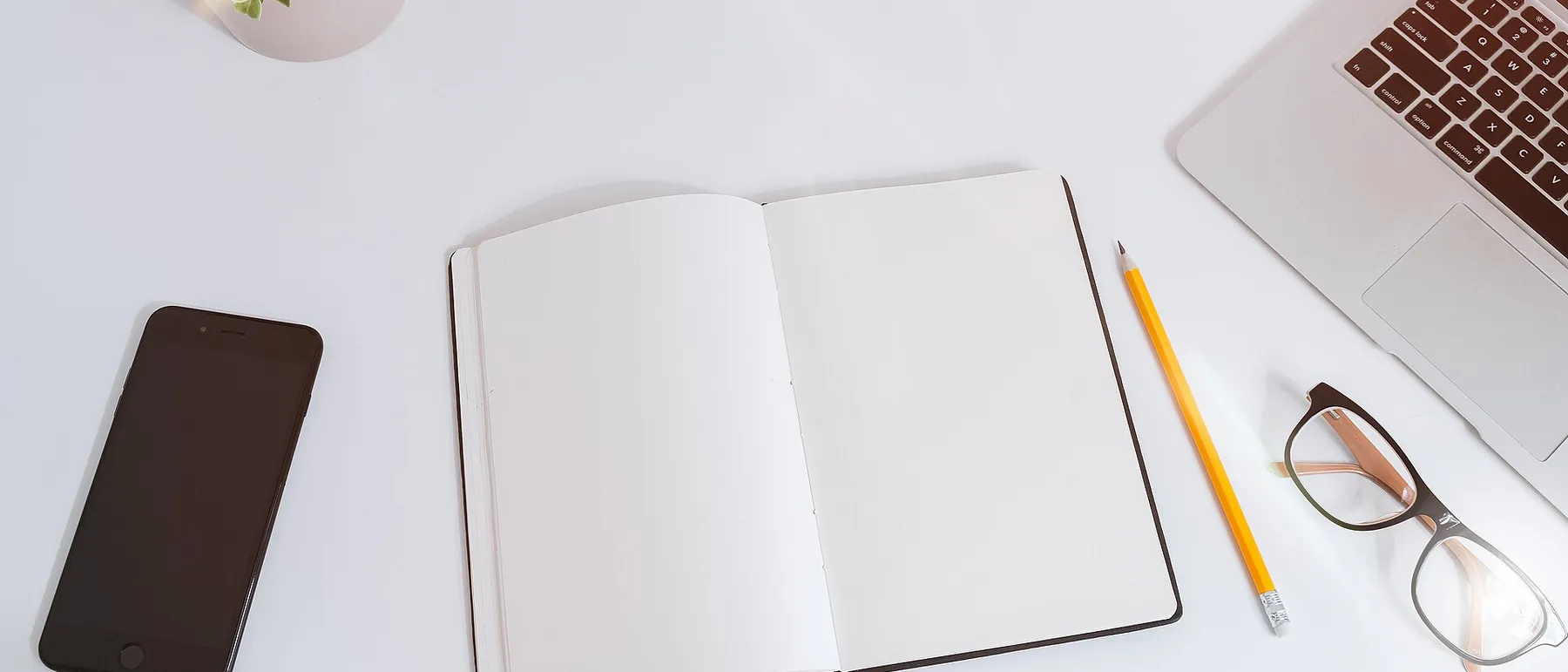 A clean white desk with an open notebook, a pencil, a phone and a laptop