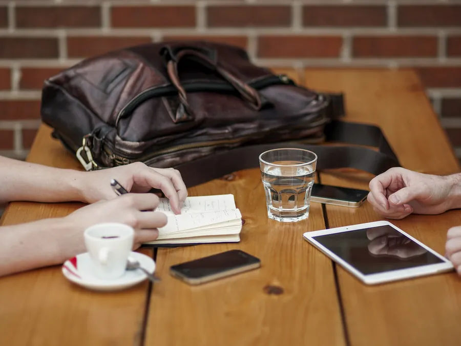 Two people talking across a wooden table with coffee, a notebook and a tablet